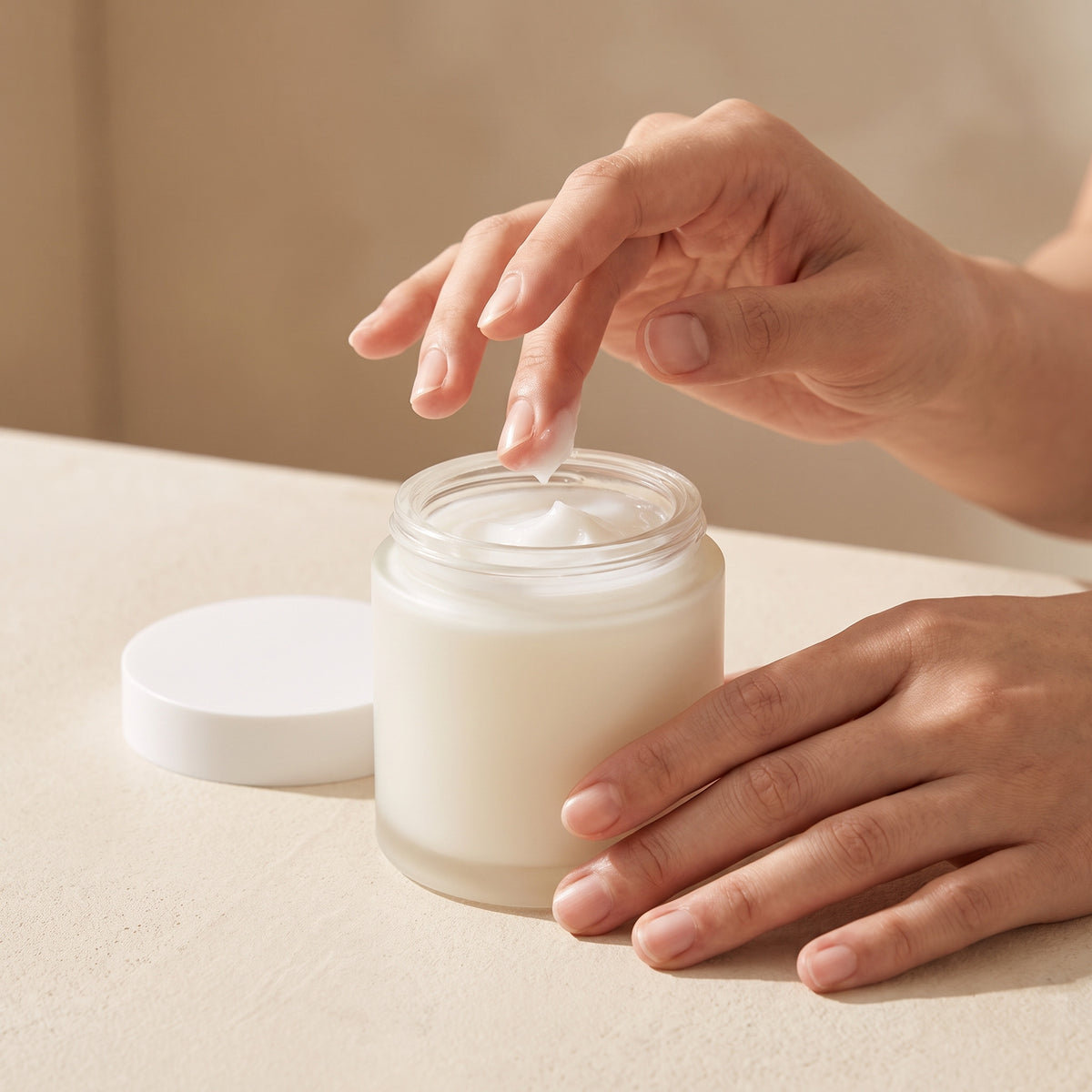 Two fingertips lifting white gel cream from an open frosted white jar standing on a pale surface